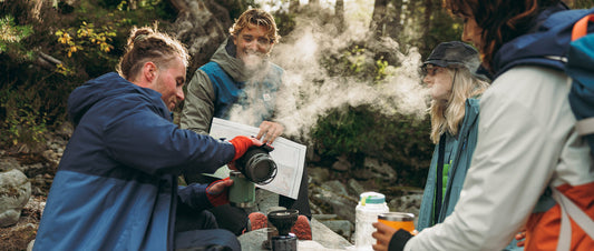 A group sit around, pouring coffee by a river