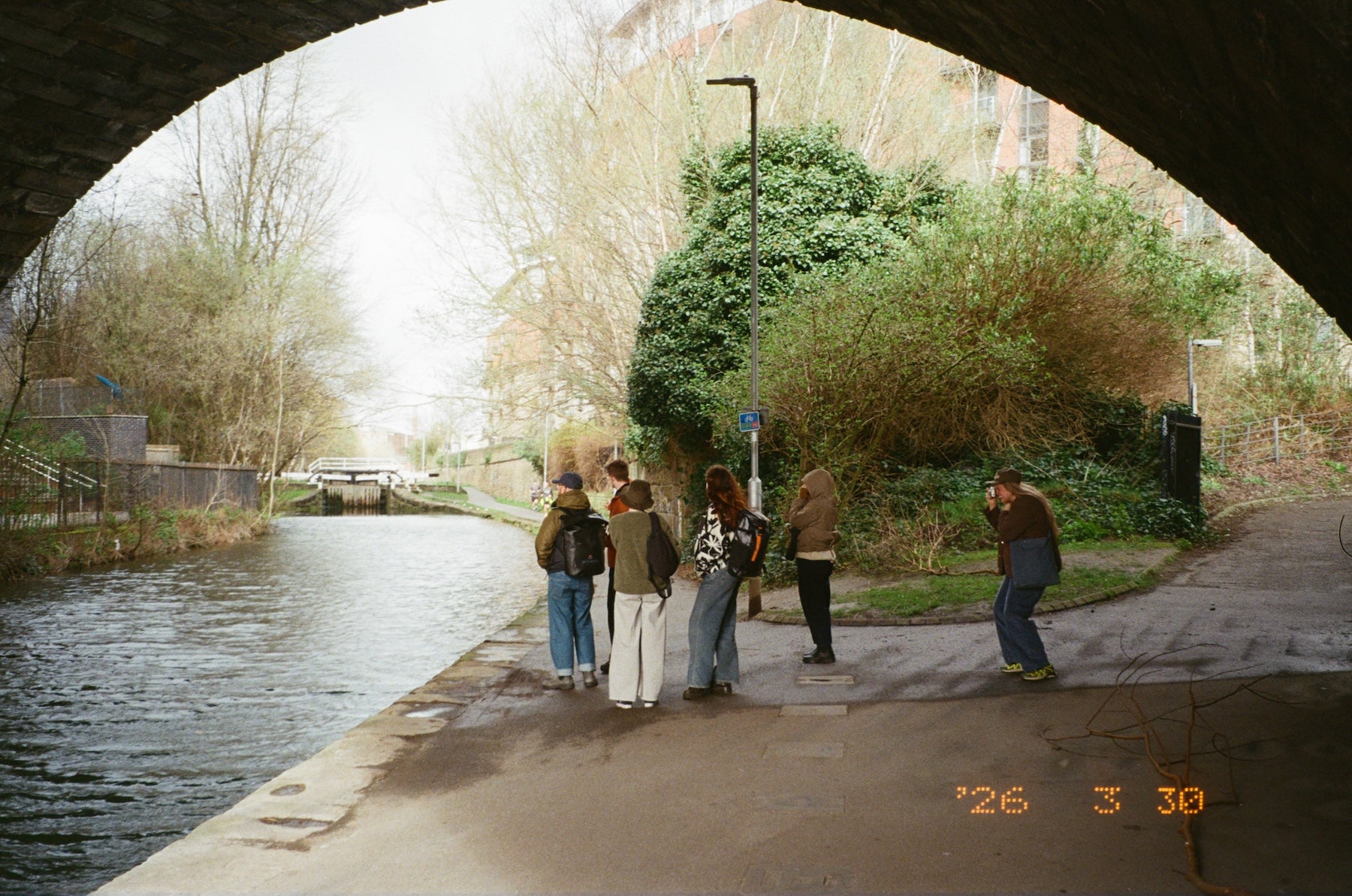 People standing by a canal under an archway with trees and buildings in the background.